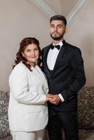 A man and woman are posing for a picture in formal attire. The man is wearing a black suit and the woman is wearing a white dress. They are both smiling and holding hands, creating a warm photo