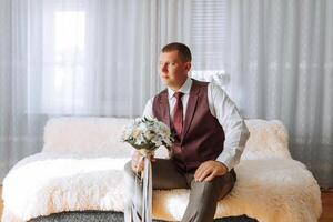 A man in a red vest sits on a bed with a bouquet of flowers in his lap photo