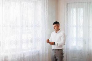 A man in a white shirt is standing in front of a window with a lace curtain. He is adjusting his tie and looking out the window. Concept of formality and attention to detail photo