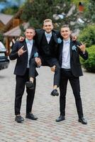 Three men in suits are posing for a picture, with one man holding the other two up. Scene is lighthearted and fun, as the men are dressed up in formal attire and are posing for a photo