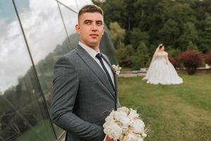 A man in a suit and tie stands in front of a glass building with a bride in a white dress behind him photo