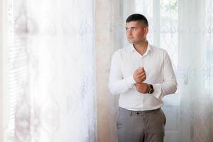 A man in a white shirt is standing in front of a window, adjusting his shirt. Concept of formality and attention to detail, as the man takes care to ensure his appearance is neat and tidy photo