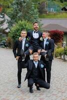 Four men in suits are posing for a picture, with one man holding the other three up. Scene is lighthearted and fun, as the men are enjoying themselves and posing for a photo together