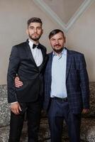 Two men in suits are posing for a picture. One of them is wearing a tie. The man on the left is wearing a black suit and the man on the right is wearing a blue suit photo