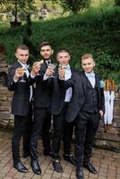 Four men in suits are posing for a photo, each holding a glass of wine. Scene is celebratory and festive, as the men are dressed in formal attire and are toasting to a special occasion photo