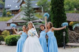 A bride and her bridesmaids are posing for a picture in front of a house. The bride is wearing a white dress and the bridesmaids are wearing blue dresses. They are all smiling and holding flowers photo