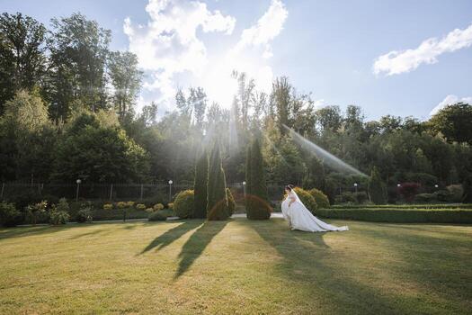 A bride is standing in a field with trees in the background. The sun is shining brightly, casting shadows on the grass. The scene is serene and peaceful, with the bride looking out over the field photo