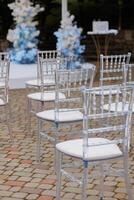 A row of chairs are set up in a courtyard, with a blue and white theme. The chairs are all clear and white, and they are arranged in a neat row. The chairs are empty photo