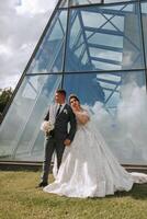 A bride and groom are standing in front of a large glass building. The bride is wearing a white dress and the groom is wearing a suit. They are holding hands and smiling at the camera photo