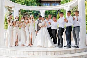 A group of people are posing for a picture, with a bride and groom in the center. The bride is wearing a white dress and the groom is wearing a suit. The group is standing in front of a white archway photo