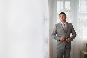 A man in a suit is standing in front of a window, adjusting his tie. Concept of formality and professionalism, as the man is dressed in a suit and tie photo