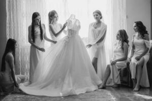 A group of women are gathered around a wedding dress, with one of them sitting on a chair. Scene is one of celebration and camaraderie photo