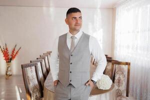 A man in a suit and vest stands in front of a long table with chairs. He is wearing a tie and he is posing for a photo. The room has a formal and elegant atmosphere, with the table photo