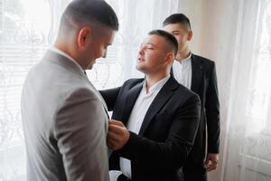 Three men are getting dressed up in suits and ties. One man is helping another man with his tie photo