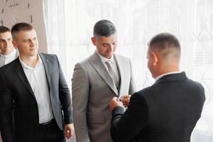 A man in a suit is getting his tie adjusted by another man. The other man is wearing a tie as well. The two men are standing in front of a window photo