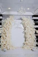 A white archway with the words written on it. The archway is decorated with white flowers and is surrounded by a white backdrop. The scene is elegant and romantic photo