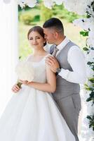 A bride and groom are posing for a picture in front of a white arch. The bride is holding a bouquet and the groom is wearing a tie. Scene is happy and romantic photo