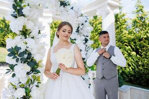 A bride and groom are standing in front of a white archway. The bride is holding a bouquet of flowers and the groom is adjusting his tie. The scene is likely a wedding ceremony or reception photo
