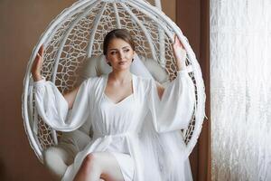 A woman in a white gown sits in a white chair. She is wearing a veil and is smiling photo