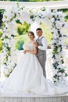 A bride and groom are posing for a picture under a white arch. The bride is holding a bouquet and the groom is wearing a vest. The scene is set in a garden or a park, with a white arch as the backdrop photo