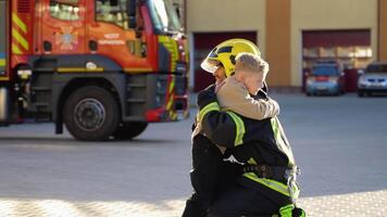 Firefighter in uniform hugs a little boy near a fire engine at station video