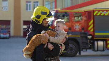 A firefighter take a little child boy to save him. Fireman with kid in his arms. Fire engine car on background video