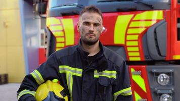 Tired firefighter in protective uniform looking at camera while standing near fire engine after extinguishing a fire video