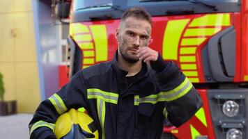 Tired fireman in protective uniform looking at camera while standing near fire engine on station video