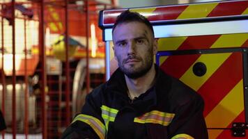 Tired fireman in protective uniform looking at camera while sitting near fire engine on station after extinguishing a fire video