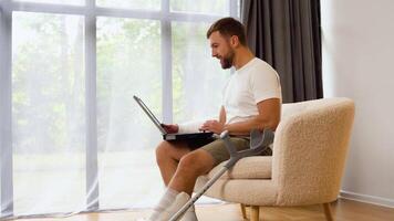 Handsome man with broken leg and arm in gypsum is using a laptop and smiling while sitting on sofa at home video