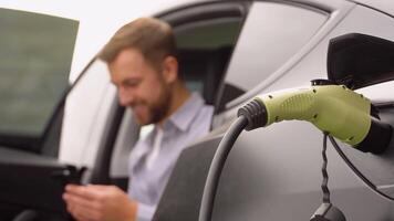 Man Charging Electric Vehicle With Cable Looking At App On Mobile Phone video
