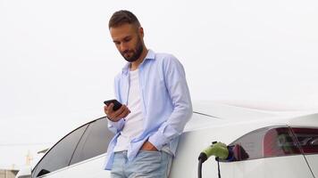 Bearded man with smartphone near electric car waiting for the finish of the battery charging process video
