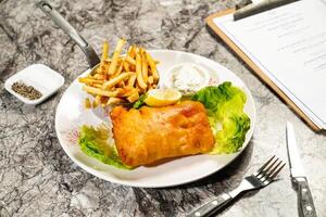 Fried cod fish and chips with tartar sauce with lemon, fork and knife served in plate isolated on marble background side view of hong kong food photo