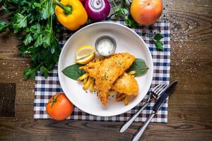 Fried Fish And Chips with lemon, dip, tomato, onion, capsicum, coriander, knife and fork served in dish isolated on napkin top view of italian fastfood on wooden table photo