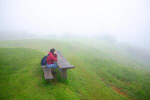 Asian young woman in red raincoat and bag sitting on wooden bench in green grass field and foggy with mist on mountain in rainy season. Travel in Asian photo