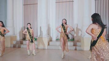 Three women in elegant pageant attire with sashes perform a dance routine in a grand hall video