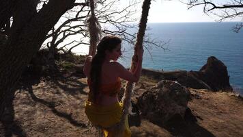 Closeup portrait of sensual young brunette woman in yellow dress, happily swinging on a rope swing outdoors isolated on blurry sea background with natural bokeh in soft warm sunset backlight. video