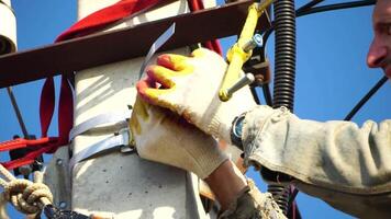 Skilled electrician in helmet fixes wires standing on ladder near high pole against blue sky on summer day backside view. Electrical service and mounting on the pole. Slow motion video