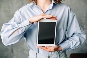 A young businesswoman is smiling as she holds a digital tablet computer, which is showing a blank, empty mock-up screen. The image is advertising software, a website or web service, and it is standing photo