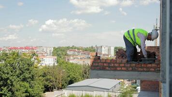 Bricklayer Working on High-Rise Construction with Scenic Background. A bricklayer, equipped with a high-visibility vest and hard hat, is diligently working on the exterior wall of a high-rise building video