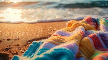 Colorful striped towel on a sandy beach near the water's edge with soft waves and a warm, glowing sunset in the background photo
