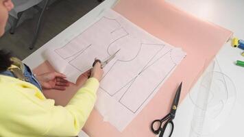 Young brunette designer is cutting a pattern for a vest with scissors on the table. Top view video
