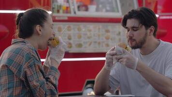 Close-up shot of a young couple of friends enjoying a burger in front of a food truck. Slow motion video
