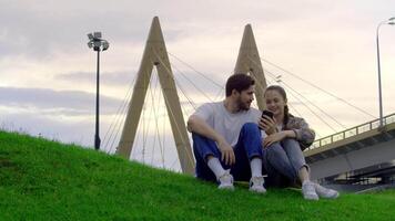 Young couple of students is sitting in a city park, enthusiastically watching something on one of their phones. Slow motion. video