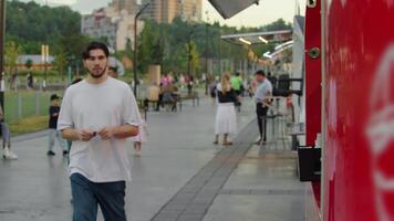 Guy walks up to the red food stand in the park and is going to get something to eat. Slow motion video