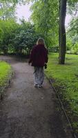 A young woman in a burgundy hoodie and patterned pants is seen from the back as she walks along a winding path in a green, leafy park. The atmosphere is calm and serene, with the overcast sky video