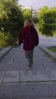 A slow-motion capturing a woman in a burgundy hoodie walking down stone steps towards a calm, reflective lake surrounded by lush greenery. The camera follows her from behind, emphasizing the video