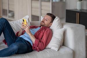 Procrastination with phone. Lazy man with social media addiction lying on couch at home scroll web photo