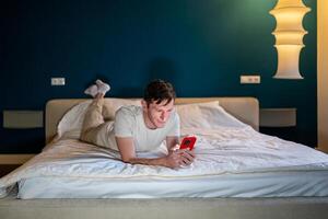 Man reading information in smartphone lying in bed at home in modern bedroom in evening. photo
