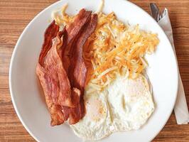 The golden-brown hash browns are piled high on a white plate with crispy bacon strips and a perfectly fried egg nestled alongside. The image evokes the comforting aroma of a classic American breakfast photo